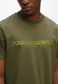Olivinės spalvos trumpų rankovių marškinėliai, pagaminti iš lygaus audinio, su ryškiu, tekstūruotu "KARL LAGERFELD" logotipu kontrastingos šviesiai žalios spalvos.