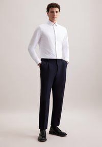 Man staat met zijn handen in zijn zakken, gekleed in een wit overhemd met lange mouwen, donkere op maat gemaakte broek en zwarte nette schoenen op een neutrale achtergrond.
