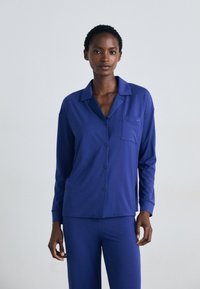 Chemise de pyjama à manches longues et à boutons, de couleur bleu foncé, en tissu doux. Comprend un col et une poche avant sur le côté gauche.