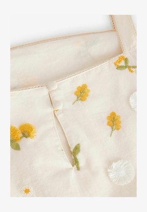 Broderad tygblus i ljusbeige med gula blommor och gröna blad, med två tygklädda knappar framtill och en delikat kant.