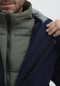 Navy outer jacket with a green insulated inner layer, featuring a high collar and dual zippers. Textured fabric with visible stitching details.