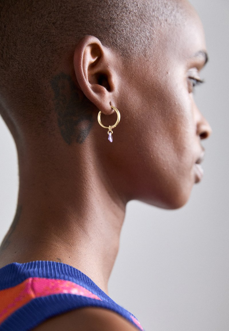 Χρυσό σκουλαρίκι σε σχήμα δαχτυλιδιού με κρεμαστό μωβ πολύτιμο λίθο, εμφανίζεται σε άτομο με κοντοκομμένο χτένισμα σε ουδέτερο φόντο.