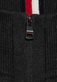 Maglione zip-up grigio scuro lavorato a maglia con texture a coste, dotato di una cerniera in metallo e un accento striato di rosso, bianco e blu marine vicino al colletto.