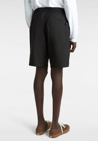 Schwarze Shorts mit einem elastischen Bund, ausgestattet mit einer kleinen, praktischen hinteren Reißverschlusstasche. Kombiniert mit braunen Schuhen, die eine glatte Textur aufweisen.