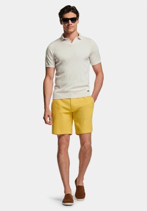 Homme portant des lunettes de soleil noires, un polo blanc à manches courtes, un short jaune et des chaussures marron sans lacets, debout devant un fond blanc.