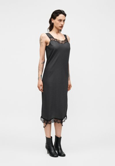 Γυναίκα με τατουάζ που φοράει μαύρο μίντι φόρεμα χωρίς μανίκια με δαντέλα και μαύρα μποτάκια, στέκεται μπροστά σε ένα απλό φωτεινό φόντο.