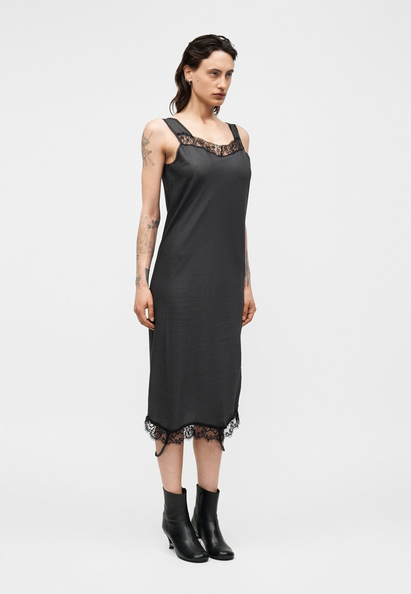 Frau mit Tattoos, die ein ärmelloses schwarzes Midikleid mit Spitzenbesatz und schwarze Stiefeletten trägt, stehend vor einem schlichten hellen Hintergrund.