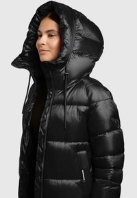 Kobieta w błyszczącej czarnej kurtce puchowej z dużym kapturem i widocznym zamkiem, zwrócona w lewo na jednolitym tle.