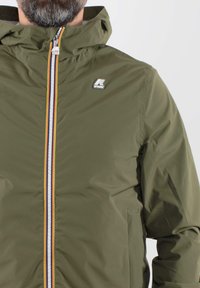 Olivgrüne Kapuzenjacke aus glattem, leichtem Material. Verfügbar mit einem Frontreißverschluss, der einen mehrfarbigen Streifen und ein Logo auf der Brust hat.