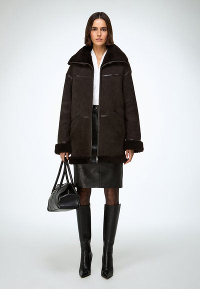 Veste en peau retournée noire avec un large col, des fermetures éclair frontales et deux poches. Associée à une jupe en cuir noire et des bottes noires montant jusqu'aux genoux.