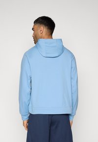 Sweat à capuche bleu clair avec cordon de serrage, manches longues et poignets côtelés. Tissu lisse sans motifs, porté avec un short bleu marine.
