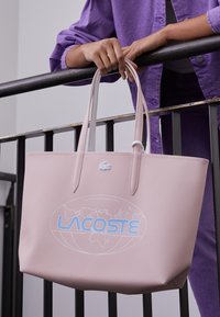 Sac fourre-tout texturé rose avec deux poignées, présentant un graphique de carte du monde bleue et le logo "LACOSTE". Matériau lisse, design moderne.