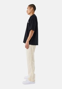 T-shirt nera in cotone con vestibilità rilassata, abbinata a pantaloni color crema con dettagli a frange sul lato e sneakers bianche.