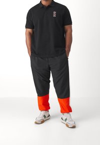 Black polo shirt with logo details and black pants featuring orange cuffs. Paired with white sneakers with green accents.
