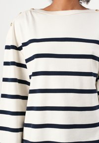 Marineblå og kremstripet topp laget av myk stoff. Har en bred halslinning og gullknappdetaljer på skuldrene. Løs passform med lange ermer.