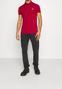 Polo shirt en coton rouge avec un col et trois boutons noirs. Présente un petit logo bleu sur la poitrine. Associé à un jean noir et des bottes.