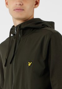Felpa con zip di colore verde scuro, dotata di cappuccio regolabile con cordino e un piccolo logo ricamato giallo sul petto. Tessuto con texture liscia.