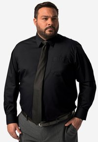 Camisetta nera a maniche lunghe con taschino, abbinata a una cravatta scura e pantaloni grigi. Il tessuto appare liscio e presenta un design formale.