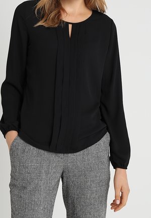 Vrouw draagt een zwart blouse met lange mouwen en een halslijn met keyhole-effect, en een grijze geruite broek, staat met één hand in haar zak.