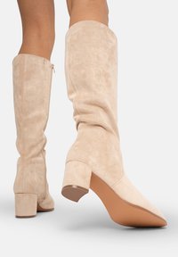 Botas de ante beige hasta la rodilla con un tacón grueso, que presentan una textura suave y un diseño de puntera ligeramente puntiaguda.