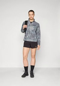 Ζακέτα Patagonia με φερμουάρ σε γκρι καμουφλάζ με στερεό γκρι τμήμα στον ώμο, συνδυασμένη με μαύρο σορτς. Η στολή περιλαμβάνει μαύρες κάλτσες και παπούτσια.