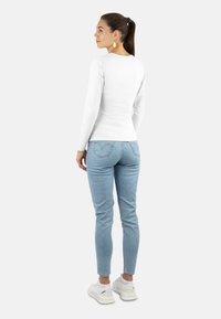 Weißes Langarmshirt kombiniert mit hellblauen Skinny Jeans und weißen Sneakers. Haare zurückgebunden, leicht zur Seite gedreht, einfache Ohrringe sichtbar.