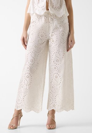 Białe spodnie o szerokich nogawkach z haftem kwiatowym z oczkami, z falistymi brzegami i sznurkiem w talii, noszone z złotymi sandałami na wysokim obcasie z paskami.