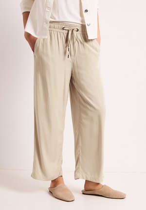 Pantalon beige à jambes larges avec taille élastique et cordon, porté avec des chaussures beige clair sans lacets et une chemise blanche partiellement visible ainsi qu'une veste.