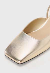Slingback flat in metallo dorato con punta quadrata, texture liscia e design minimalista. Presenta un tacco basso e un cinturino curvo alla caviglia.