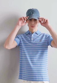 Ανοιχτό μπλε πόλο μπλουζάκι με λευκές ρίγες, κοντά μανίκια και γιακά. Ένα τζιν καπέλο είναι τοποθετημένο πάνω στο κεφάλι του μοντέλου, με ένα λογότυπο μπροστά.