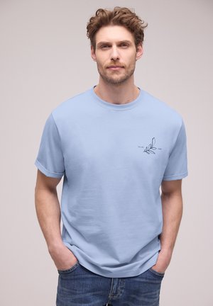 Mężczyzna z kręconymi włosami, ubrany w jasnoniebieską koszulkę z małym wzorem liścia i cyframi na lewej piersi, ręce w kieszeniach jeansów, neutralne tło.