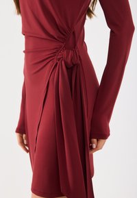 Vestido bordeaux feito de um tecido elástico, com mangas longas, franzidos laterais e um detalhe amarrado no quadril, criando um efeito drapeado.