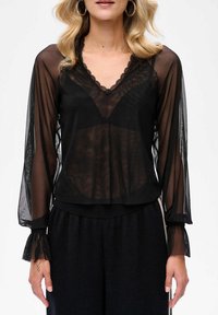 Czarna, przezroczysta bluzka z długimi, luźnymi rękawami, z dekoltem w kształcie V z falbanką oraz teksturowanym wzorem. Połączona z czarnymi spodniami na elastycznej talii.
