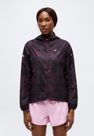 Femme portant une veste zippée noire et violette à motifs avec capuche et un short de sport rose clair, debout devant un fond uni.