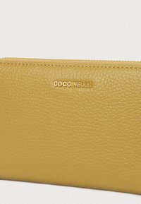 Portofel galben din piele texturată, cu fermoar auriu și logo-ul auriu "COCCINELLE" centrat pe partea frontală.