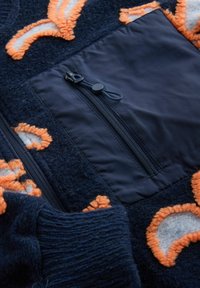Marineblaue Fleecejacke mit orangefarbenen und grauen, strukturierten Halbmondmustern sowie einer schwarzen Reißverschlusstasche auf der Brust.