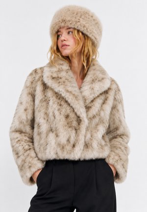 Femme portant une veste en fausse fourrure beige et un chapeau assorti, les mains dans les poches d’un pantalon noir, regardant légèrement vers sa gauche.
