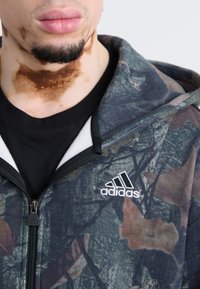Hooded zip jacket in camouflage pattern with shades of green, brown, and gray. Features an embroidered white logo on the chest and zipper detail.