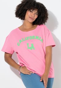 Růžové bavlněné tričko s krátkým rukávem, na kterém je zelený vyšitý nápis "CALIFORNIA LA" přes přední část. Volný střih, kulatý výstřih.