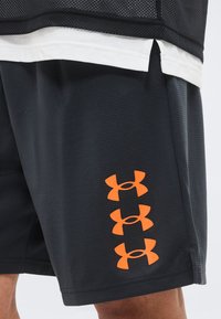 Μαύρα αθλητικά σορτς με τρία κάθετα πορτοκαλί λογότυπα Under Armour στο αριστερό μηρό, φορεμένα με στρωματοποιημένα μαύρα και λευκά μπλουζάκια.