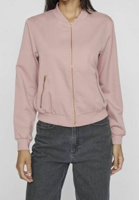 Femme portant une veste bomber zippée rose clair avec col, poignets et ourlet côtelés, associée à un jean taille haute gris foncé.