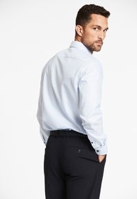 Lichtblauw overhemd met lange mouwen en een gestructureerde kraag, gecombineerd met een donker broek. Het overhemd heeft subtiele strepen en knopen op de manchetten.