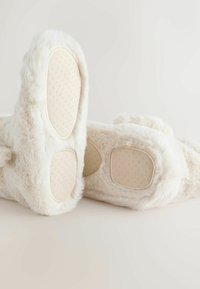 Papuci din blană artificială de culoare crem, cu o talpă texturată de nuanță maronie. Formă rotundă, exterior moale și brant căptușit pentru confort.
