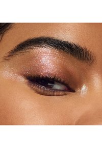 Gros plan sur un œil avec un fard à paupières rose scintillant, des cils noirs épais et des sourcils bien entretenus. La peau présente une texture lisse et éclatante.