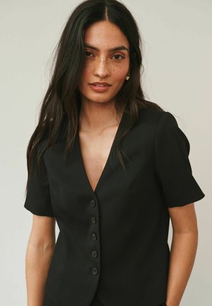 Woman with long dark hair and freckles wearing a black short-sleeve buttoned blazer, facing forward with a neutral expression.