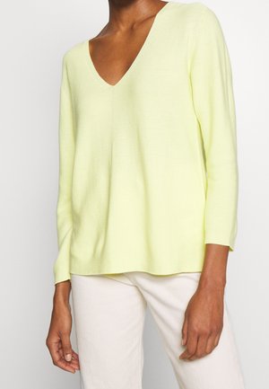 Osoba ubrana w jasnobeżowy sweter z długim rękawem o dekolcie w serek oraz białe spodnie, stojąca na tle jednolitego białego tła.