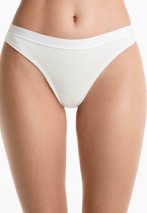 Witte katoenen stringonderbroek wordt tentoongesteld, met een merkband rond de taille. De focus ligt op de onderbuik en de bovenbenen.