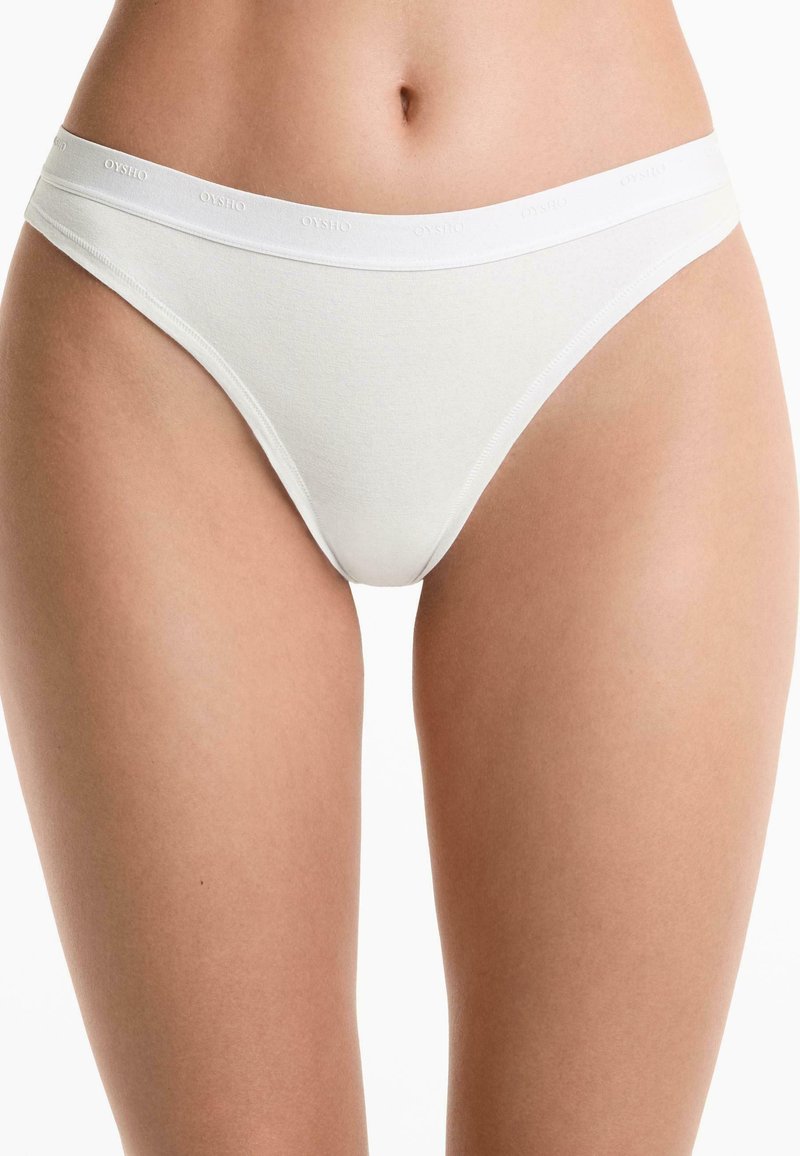 Weiße Baumwoll-Tanga-Unterwäsche wird präsentiert, mit einem gebrandeten Bund. Der Fokus liegt auf dem unteren Torso und den oberen Oberschenkeln.