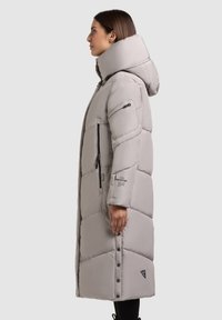 Kvinde i en beige lang polstret frakke med høj krave, lynlåslommer og trykknapper langs siden, stående i profil mod en ensfarvet baggrund.