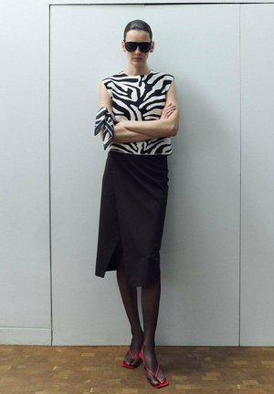 Frau mit ärmellosem Oberteil mit Zebra-Muster, schwarzem Rock mit Schlitz, durchsichtigen schwarzen Strumpfhosen, roten Sandalen mit Absatz und schwarzen Sonnenbrillen, stehend mit verschränkten Armen.